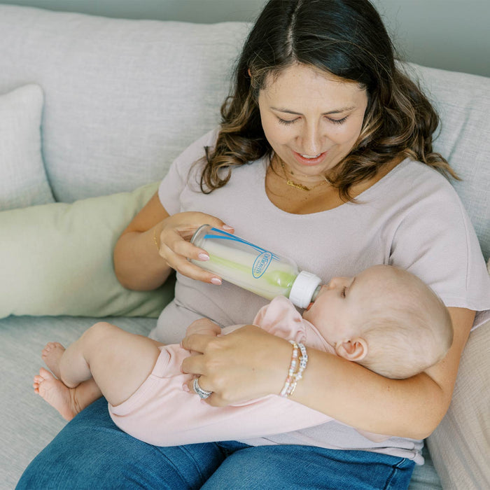 A woman sitting on a light gray couch feeds a baby from a glass bottle with a white cap. The woman wears a light pink t-shirt and blue jeans, and the baby wears a pale pink outfit.