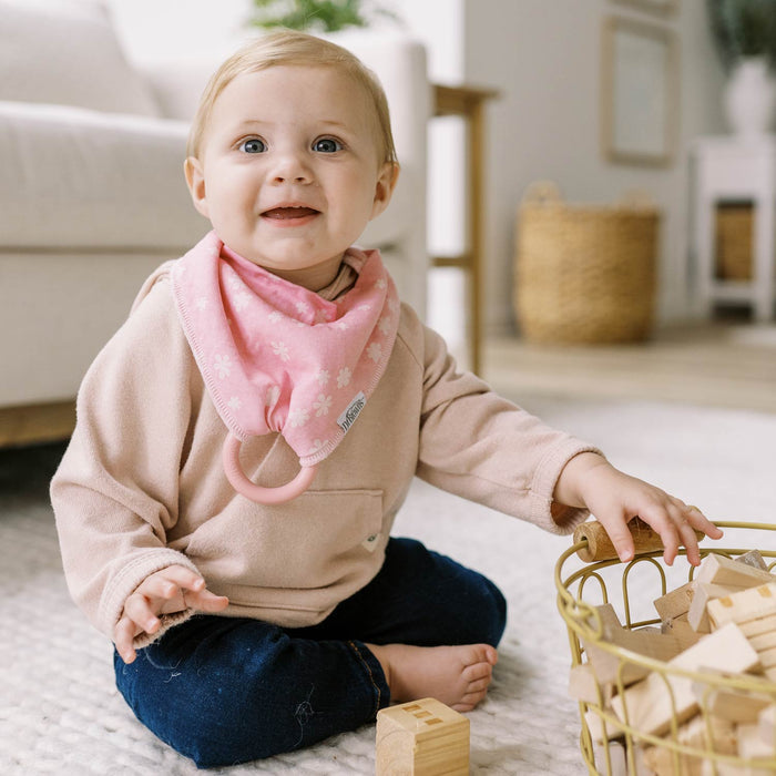 A smiling baby sits on the floor playing with wooden blocks, wearing a light sweater and Dr. Brown's™ Bandana Bibs with Removable Teether by Dr. Brown's, next to a wire basket in a cozy, well-lit room.