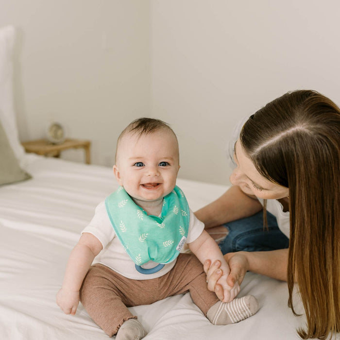 A teething baby smiles on a bed, wearing Dr. Brown's™ Bandana Bibs with Removable Teether, holding hands with a woman. Soft natural light and neutral decor create a cozy scene.