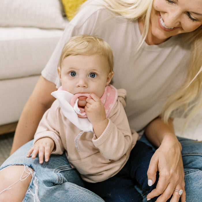 A smiling woman sits on the floor with a blond baby in her lap. The baby, wearing Dr. Brown's™ Bandana Bibs with Removable Teether, looks up and chews on the teether while the woman gently holds her.
