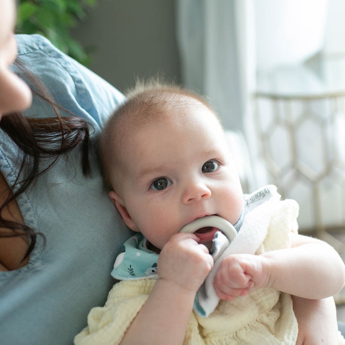 A light-haired baby with big eyes sits on an adult’s lap, wearing Dr. Brown's™ Bandana Bibs with Removable Teether and wrapped in a yellow blanket. The bib is super-absorbent and features a removable teether. The adult’s face is partially out of frame.