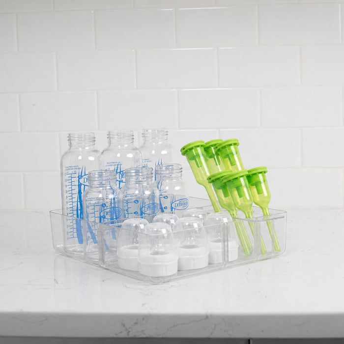 A clear plastic organizer tray holding several transparent baby bottles with blue measurement markings and a set of bright green breast pump tubes, placed on a white countertop against a white subway tile wall.