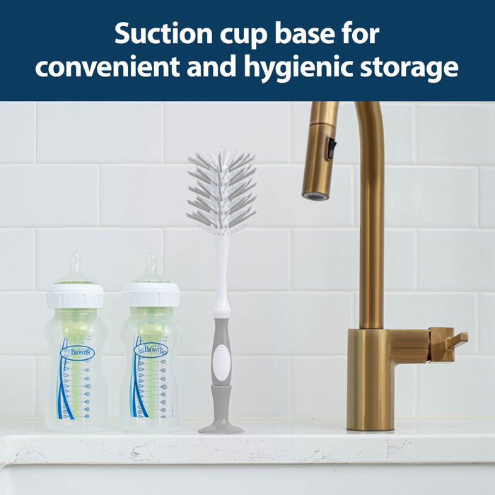 The Dr. Brown’s™ Deluxe Bottle Brush with a suction cup base stands upright for easy cleaning beside two baby bottles and a brass kitchen faucet on a white countertop.