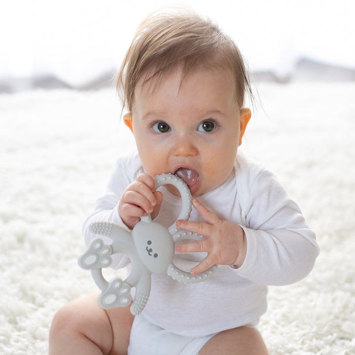 A baby in a white onesie sits on a soft white surface, chewing on Dr. Brown's® Flexees™ Bunny Silicone Teether to help soothe teething discomfort.