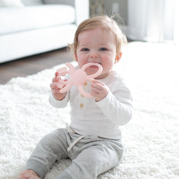 A baby sits on a white carpet, chewing on Dr. Brown's® Flexees™ Bunny Silicone Teether, with a white sofa and curtains in the background—a sweet teething moment with Dr. Brown's.