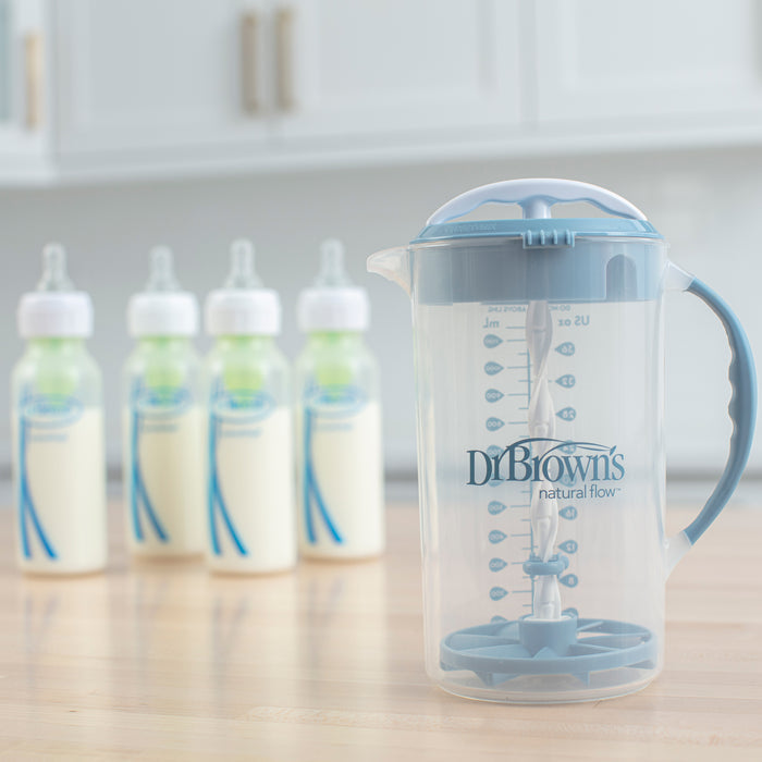 A Dr. Brown's® Formula Mixing Pitcher by Dr. Brown's with a blue handle rests on the countertop, ideal for mixing multiple servings of baby formula, with four filled bottles blurred in the background.