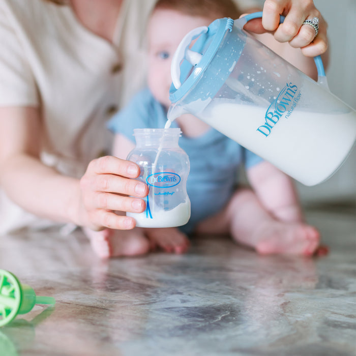 An adult pours milk from a Dr. Brown's® Formula Mixing Pitcher into a Dr. Brown's baby bottle, while a baby in blue sits on the floor in the background.