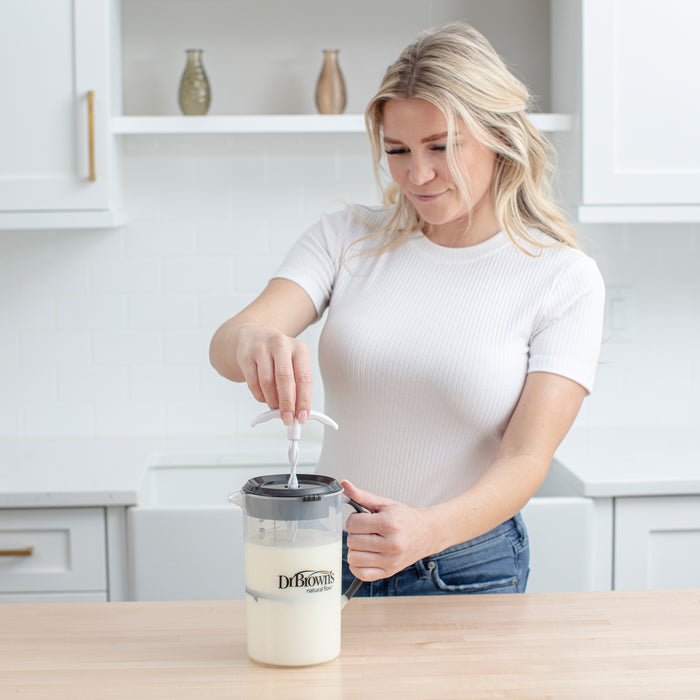 A woman uses a Dr. Brown's® Formula Mixing Pitcher on a wooden countertop to blend formula, making baby bottle prep easy and helping reduce gas.