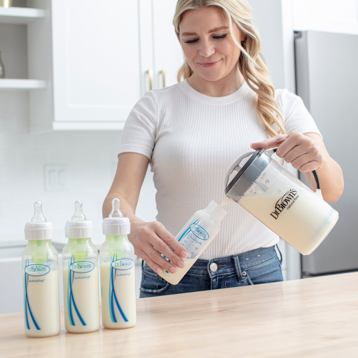 A woman uses the Dr. Brown's® Formula Mixing Pitcher by Dr. Brown's to pour formula into a baby bottle on her kitchen counter, with three filled bottles beside her, designed to help reduce gas for babies.