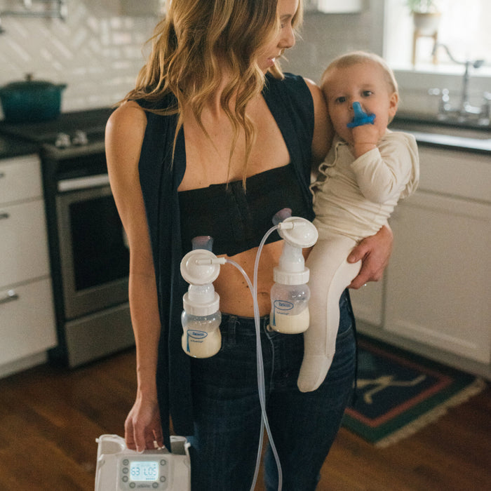 A woman stands in a kitchen holding a baby, effortlessly using an electric pump with two bottles partially filled with milk. Thanks to her Dr. Brown's Hands Free Pumping Bra from Dr. Brown's, she enjoys a custom fit while the baby, dressed in white, plays with a blue toy.