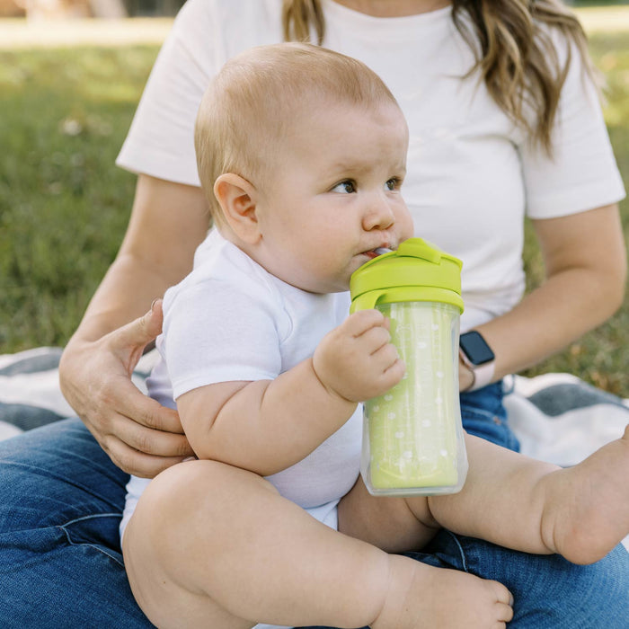 A baby in a white onesie sits on an adult’s lap outdoors, sipping from Dr. Brown’s® Milestones™ Insulated Straw Cup, 2-Pack. They’re on a grassy blanket, enjoying the spill-proof cup; the adult’s face isn’t visible.