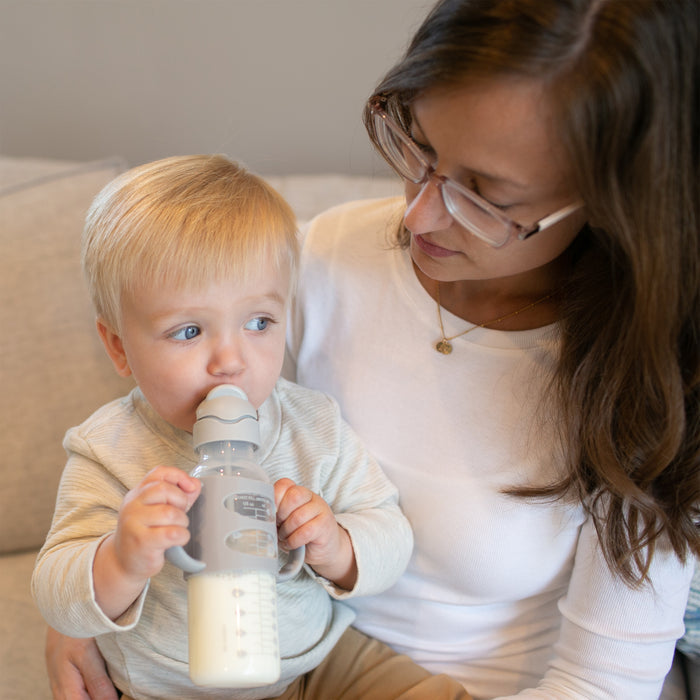 A woman sits on a couch holding a young child who is drinking milk from Dr. Brown's Milestones Narrow Sippy Straw Bottle with Silicone Handles, 8 oz/250 mL, showcasing early baby drinking skills.