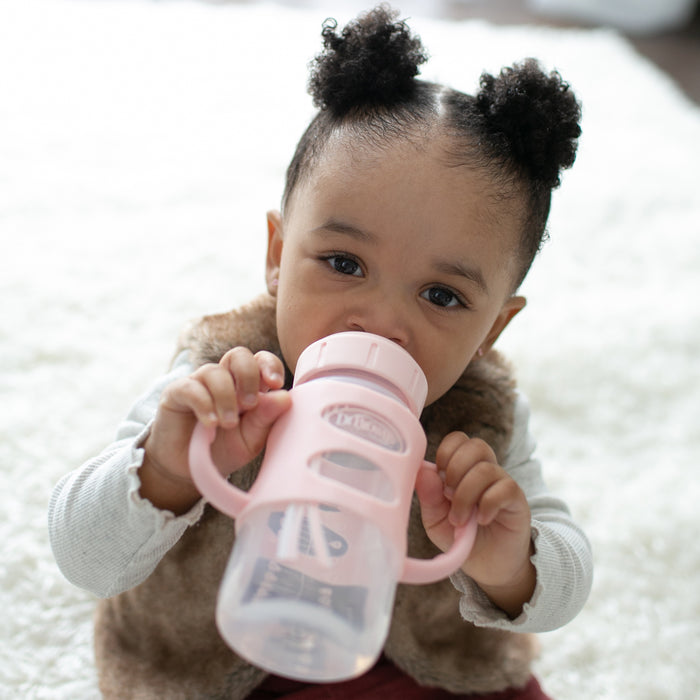 A toddler with two puffs in her hair sits on a white rug, drinking from Dr. Brown's Milestones Wide-Neck Sippy Spout Bottle with Silicone Handles, 9 oz, in pink.