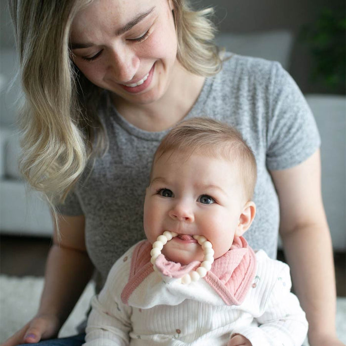 A woman smiles at a baby on her lap. The baby wears a Dr. Brown’s® Muslin Bandana Bib with a Flexees™ Beaded Teether Ring, soothing their teething gums while looking adorable in the trusted comfort of Dr. Brown’s.