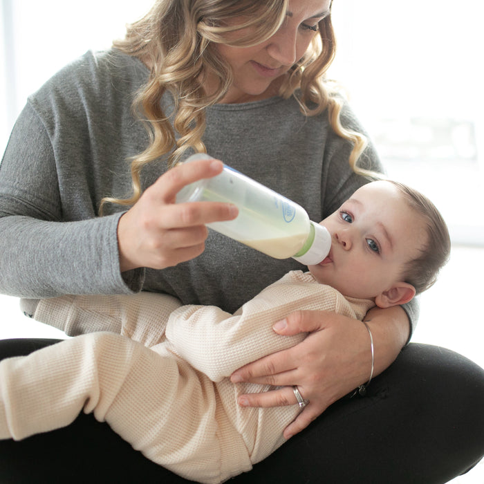 A woman holds a baby on her lap and feeds them with a Dr. Brown’s Natural Flow® Anti-Colic Options+™ Narrow Baby Bottle with Level 1 Slow Flow Nipple, 8 oz/250 mL. The baby, dressed in a light-colored outfit, looks at the camera while enjoying their pediatrician recommended Dr. Brown’s bottle.