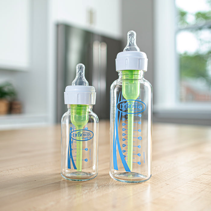 Two clear Dr. Brown’s Natural Flow® Anti-Colic Options+™ Narrow Baby Bottles with Level 1 Slow Flow Nipples, each holding 8 oz/250 mL, stand on a wooden kitchen counter, with a blurred refrigerator and window in the background. Pediatrician recommended, these Dr. Brown’s bottles are designed to provide an anti-colic feeding experience for your little one’s comfort.