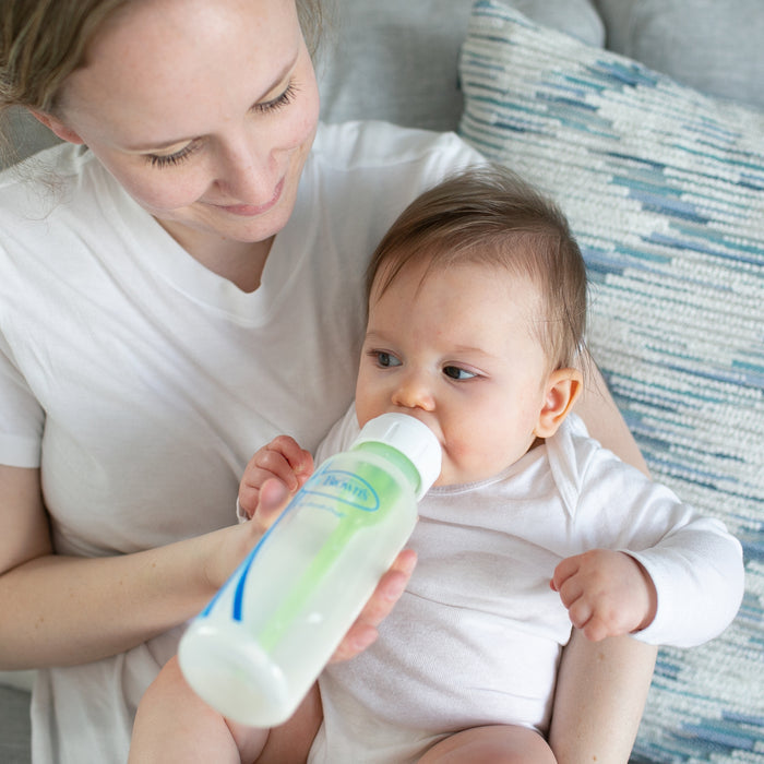 An adult in a white shirt feeds an infant in a white onesie with a Dr. Brown’s Natural Flow® Anti-Colic Options+™ Narrow Baby Bottle with Level 1 Slow Flow Nipple, 8 oz/250 mL, while sitting on a couch with a patterned pillow.
