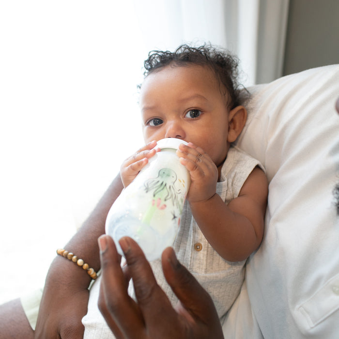 A baby dressed in white sits on an adult’s lap, holding and drinking from a Dr. Brown’s Natural Flow® Anti-Colic Options+™ Wide-Neck Baby Bottle with Level 1 Slow Flow Nipple, 9 oz/270 mL. This pediatrician-recommended bottle from Dr. Brown’s helps make feeding more comfortable for your little one.