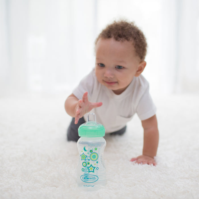 A baby in a white shirt crawls on a white carpet, reaching for a green and clear Dr. Brown’s Natural Flow Anti-Colic Options+ Wide-Neck Sippy Bottle Starter Kit with star and moon designs.