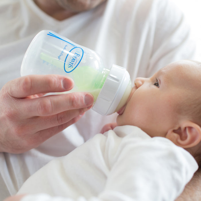 An adult feeds a baby in white clothing using Dr. Brown's Natural Flow Wide-Neck Baby Bottle with a silicone nipple, filled with light green liquid, while the baby lies comfortably during the feeding.