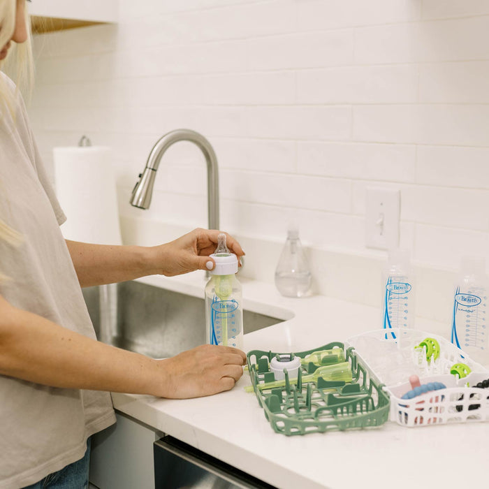 A person uses Dr. Brown's™ Options+™ Dishwasher Basket for Narrow Baby Bottle Parts at the kitchen sink, with clean bottles and a drying rack on the white countertop, creating a bright, organized space for easy cleaning.