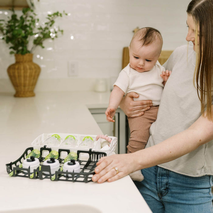 A woman holding a baby places Dr. Brown's™ Options+™ Dishwasher Basket for Narrow Baby Bottle Parts on the kitchen counter, preparing it for cleaning. A vase with greenery sits in the background.
