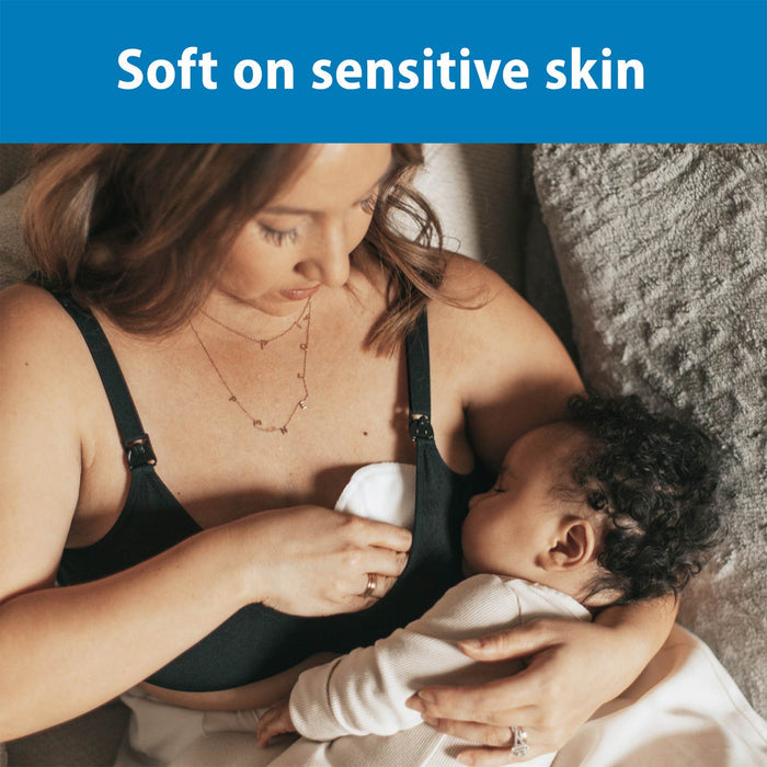 A woman breastfeeds her baby while holding Dr. Brown's™ Reusable Breast Pads to her chest. Both relax on a couch under a cozy blanket. Text above reads: "Soft on sensitive skin.
