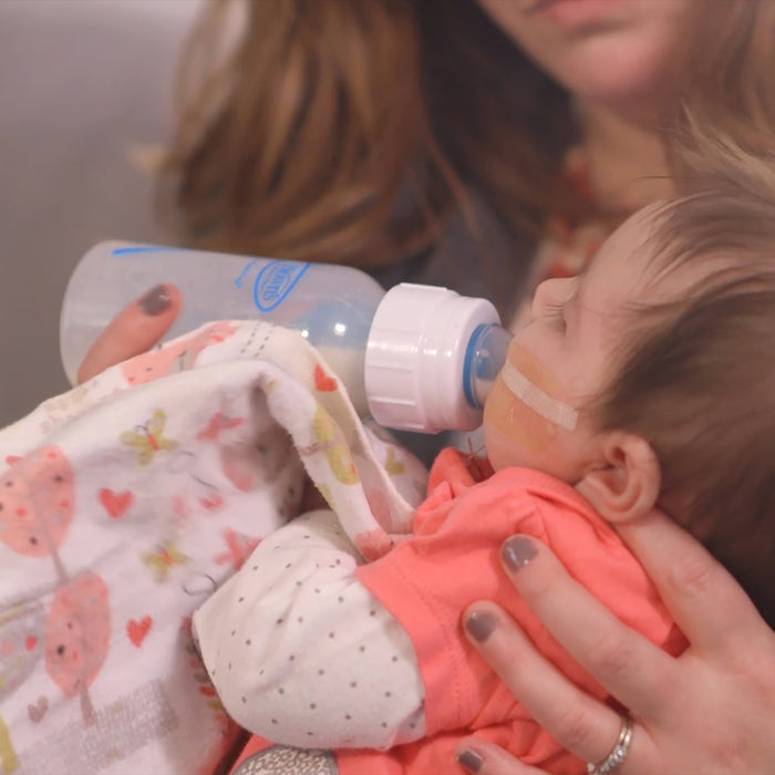 An adult feeds a baby, who has a feeding tube and is wrapped in a patterned blanket, using the Dr. Brown's Specialty Feeding System, 2 Count by Dr. Brown's—designed to assist infants with oral feeding challenges.